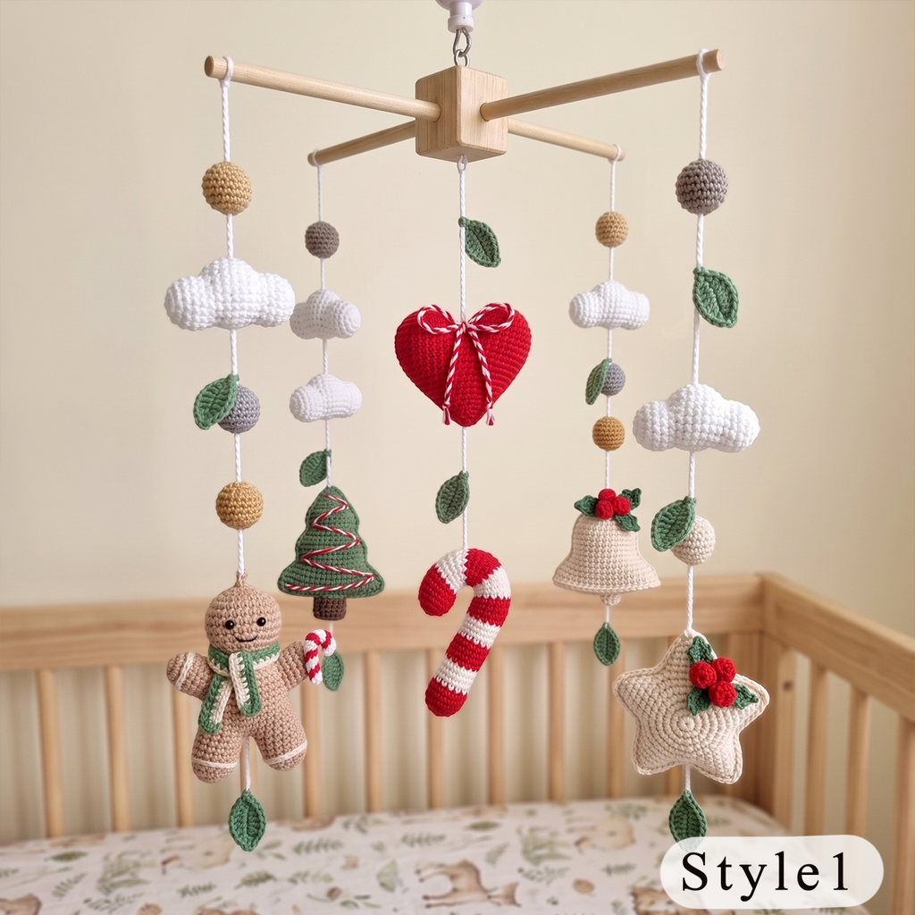 A handmade crochet baby mobile hanging over a wooden crib. The mobile features a light wood crossbar, white yarn clouds, a red crocheted heart with a bow, green leaves, candy canes, a gingerbread man, a Christmas tree, a bell, and a star with a red berry sprig.